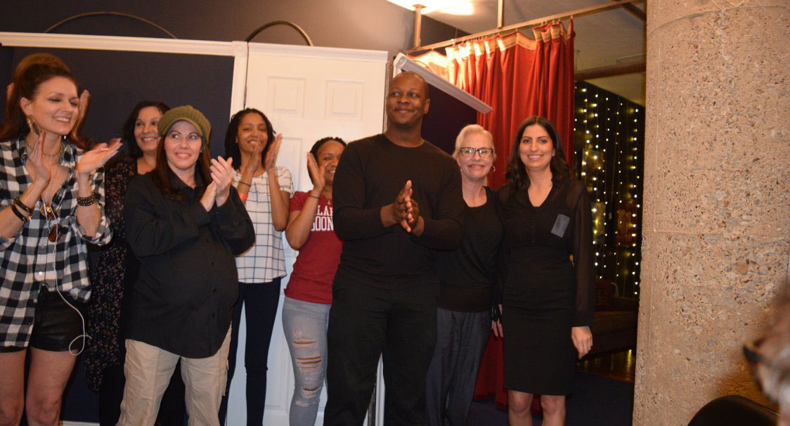 A group of six people clapping and smiling indoors.