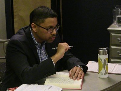 Man in glasses writing in a notebook while holding a pen.