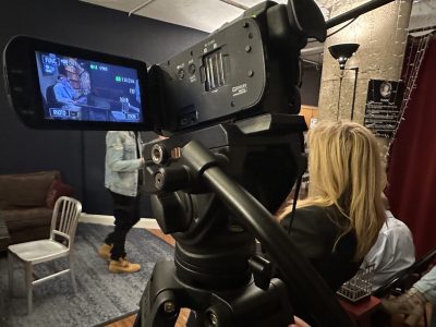 A woman operates a professional video camera filming a man speaking indoors.