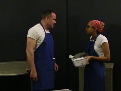 Two people in aprons talking in a kitchen or workspace.