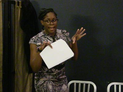 Woman speaking and holding a piece of paper in front of a chair.