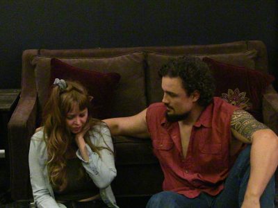 A man comforts a sad woman on a couch in a dimly lit room.